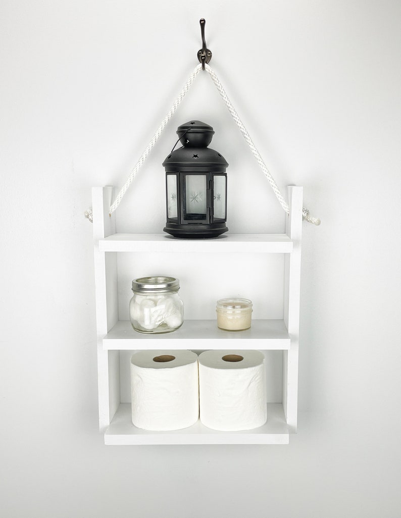 May include: White wooden shelf with two shelves, hanging from a hook on a white wall. The top shelf has a black lantern with a white star design. The middle shelf has a clear glass jar with white cotton balls and a small white candle. The bottom shelf has two rolls of white toilet paper.