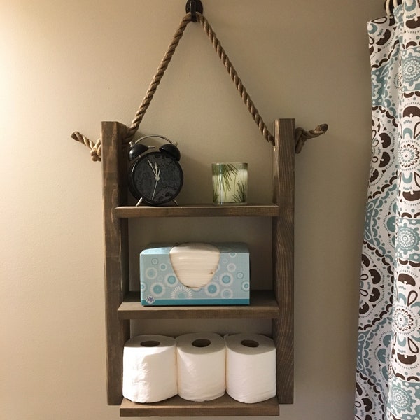 May include: A rustic wooden shelf with three tiers, hanging from a rope and a hook. The shelf is decorated with a clock, a candle, a box of tissues, and three rolls of toilet paper.