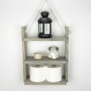 May include: A gray wooden shelf with three tiers, hanging from a white rope and a black hook. The top tier has a black lantern, the middle tier has a small candle and a glass jar with cotton balls, and the bottom tier has two rolls of toilet paper.