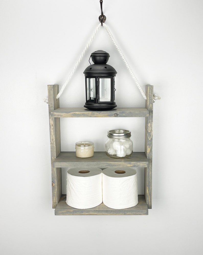 May include: A gray wooden shelf with three tiers, hanging from a white rope and a black hook. The top tier has a black lantern, the middle tier has a small candle and a glass jar with cotton balls, and the bottom tier has two rolls of toilet paper.