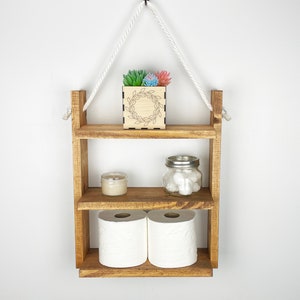 May include: A rustic wooden hanging shelf with three tiers. The top tier has a small wooden box with a wreath design and a succulent plant. The middle tier has a small candle and a glass jar with cotton balls. The bottom tier has two rolls of toilet paper. The shelf is hanging from a white rope.
