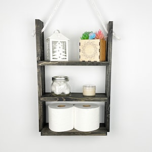 May include: A three-tiered wooden shelf with a rope hanger. The shelf is painted black and has a rustic finish. The top shelf has a white lantern with a snowflake design and a small wooden box with a wreath design. The middle shelf has a glass jar with cotton balls and a small candle. The bottom shelf has two rolls of toilet paper.