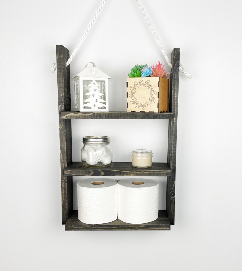 May include: A three-tiered wooden shelf with a rope hanger. The shelf is painted black and has a rustic finish. The top shelf has a white lantern with a snowflake design and a small wooden box with a wreath design. The middle shelf has a glass jar with cotton balls and a small candle. The bottom shelf has two rolls of toilet paper.