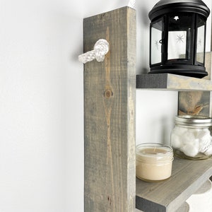 May include: A gray wooden ladder shelf with three shelves. The shelves are decorated with a candle, a jar of cotton balls, and a black lantern. The shelf is attached to the wall with a white rope.