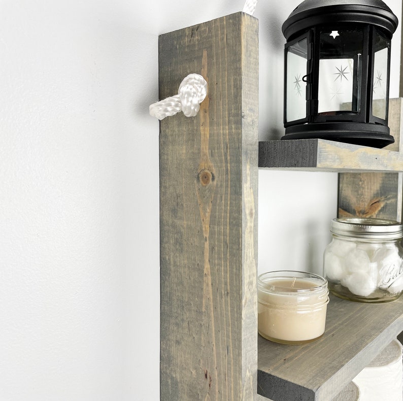 May include: A gray wooden ladder shelf with three shelves. The shelves are decorated with a candle, a jar of cotton balls, and a black lantern. The shelf is attached to the wall with a white rope.