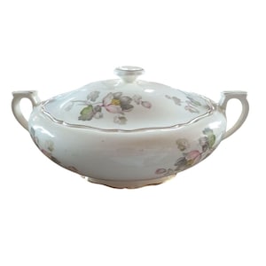 Puede incluir: Una fuente de cerámica blanca con tapa, con un estampado floral en rosa, gris y verde. La fuente tiene dos asas y un borde plateado. Una pieza clásica para servir alimentos.