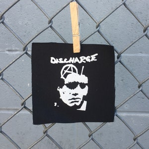 Peut inclure: Patch de groupe Discharge noir et blanc avec un symbole d'anarchie blanc et un visage de personne portant des lunettes de soleil. Le patch est accroché à une clôture en treillis métallique.