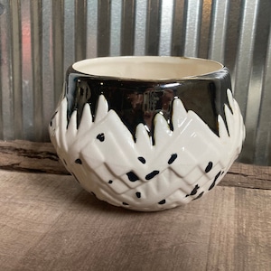 Puede incluir: Una maceta de cerámica blanca con un patrón geométrico en blanco y negro. La maceta tiene un esmalte negro en el borde superior y un acabado brillante.
