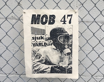 Peut inclure: Une affiche en noir et blanc avec le texte "MOB 47" et "sjuk VÄRLD" est épinglée sur une clôture en treillis métallique. L'affiche représente une personne portant un masque à gaz. L'arrière-plan est un mur de briques gris clair.