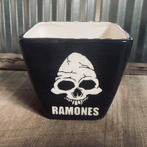 Puede incluir: Maceta cuadrada de cerámica negra con un diseño de calavera blanca y la palabra "RAMONES" en blanco. El interior es blanco. Adecuada para plantas, esta maceta es un elemento decorativo para el hogar o jardín.