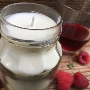 May include: A white candle in a clear glass jar with a white wick. The candle is surrounded by red raspberries on a wooden surface. There is a glass of red liquid in the background.