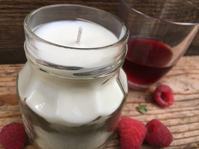 May include: A white candle in a clear glass jar with a white wick. The candle is surrounded by red raspberries on a wooden surface. There is a glass of red liquid in the background.