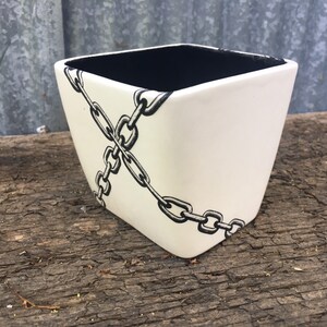 May include: A square, off-white ceramic planter with a black interior. A black and white chain design crosses the front of the planter. The planter sits on a weathered wooden surface.