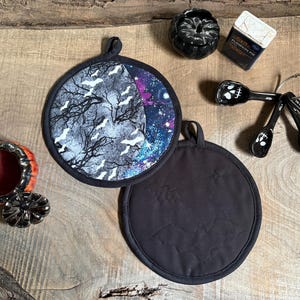 May include: Two round fabric pot holders, one featuring a black and white bat and tree pattern with a galaxy-themed side, and the other solid black with quilted bat motifs. Also includes a small pumpkin-shaped bowl, a spice tin, and measuring spoons.