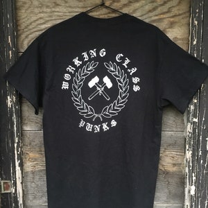 May include: Black t-shirt with a white graphic on the back. The graphic features the words "WORKING CLASS PUNKS" in a circular design with crossed hammers and a laurel wreath. The shirt is hanging on a black hanger.