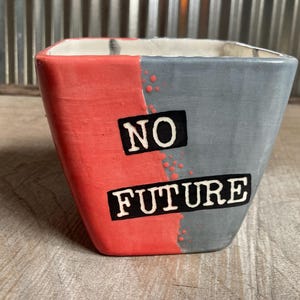 Puede incluir: Un macetero de cerámica rojo y gris con las palabras "NO FUTURE" en letras negras.