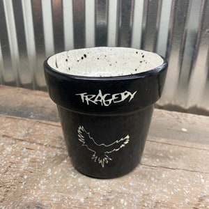 Puede incluir: Maceta de cerámica negra con motas blancas y un diseño blanco de un pájaro en vuelo. La maceta tiene la palabra "TRAGEDY" escrita en blanco en el lateral.