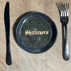 May include: A black ceramic plate with the text "Shitlickers" in gold lettering, flanked by a black knife on the left and a black fork on the right. The plate has a dark green interior and a slightly irregular edge.