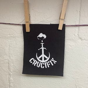 May include: A black fabric patch with a white peace sign and a sword through it. The word "CRUCIFY" is written in a stylized font below the peace sign.