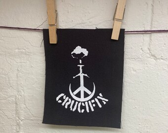 Puede incluir: Un parche de tela negro con un símbolo de paz blanco y una espada que lo atraviesa. La palabra "CRUCIFY" está escrita en una fuente estilizada debajo del símbolo de paz.