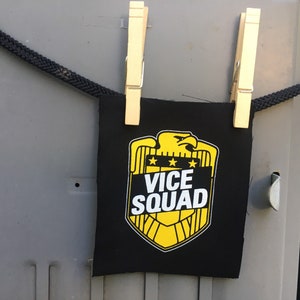 May include: A black fabric patch with a yellow eagle and shield design. The shield has the text "VICE SQUAD" in white letters.