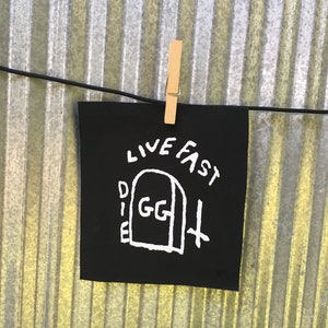 Könnte beinhalten: Ein schwarzes quadratisches Patch mit den Worten "LIVE FAST DIE GG" in weißer Schrift. Das Design beinhaltet einen Grabstein und ein Kreuz. Der Patch wird von einer Holzklammer an einer schwarzen Schnur vor einem Wellblechhintergrund gehalten.