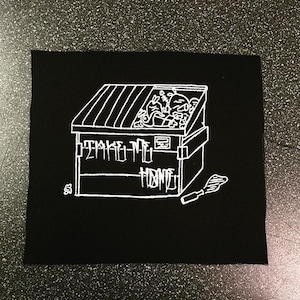 Puede incluir: Parche bordado en blanco y negro de un contenedor de basura con las palabras "Take Me Home" en el lateral. También se borda una pequeña espátula blanca en el parche.