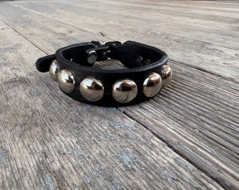 May include: A black leather bracelet featuring a row of polished, rounded silver-tone metal studs. The band is secured with a metal buckle closure, showcasing a smooth texture on the leather strap and a reflective finish on the circular hardware.