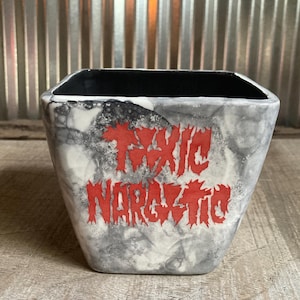 Puede incluir: Una maceta cuadrada de cerámica con un diseño veteado gris y blanco. La maceta presenta las palabras "TOXIC NARCOTIC" en rojo, con letras estilizadas. El interior de la maceta es negro.