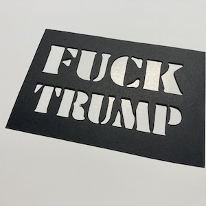 May include: Black stencil with the words "FUCK TRUMP" cut out in bold, white capital letters. The stencil is rectangular and angled, set against a white background.