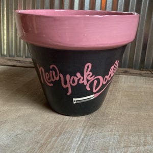May include: A black and pink painted flower pot with the words "New York Dodgers" in pink script. The rim of the pot is pink, and the pot is sitting on a wooden surface.