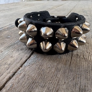 Op de afbeelding: Een zwarte leren polsband met twee horizontale rijen gepolijste zilverkleurige metalen kegelvormige studs. De armband is voorzien van een metalen gespsluiting en heeft een gestructureerd leren oppervlak met uniforme metalen details.