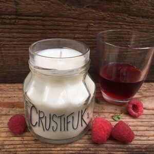 May include: A white candle in a clear glass jar with the word "CRUSTIFUK" printed on the front. The candle is surrounded by raspberries and a glass of red wine.