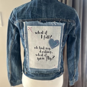 May include: A blue denim jacket with a rectangular patch on the back featuring the text "what if I fall? oh but my darling, what if you fly?" A blue heart is on the patch. The jacket is displayed on a mannequin.
