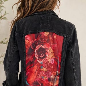 May include: Black denim jacket with a vibrant floral design on the back. The artwork features large red roses, feathers, and other flowers in shades of red, pink, and yellow. The jacket has a classic button-up design.