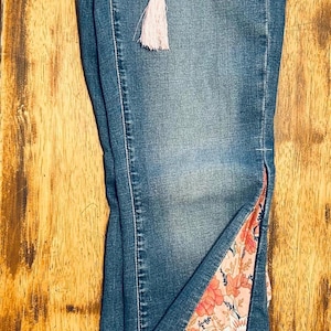 May include: Blue denim jeans with a floral patterned fabric insert on the outer seam. The jeans have a decorative pink tassel hanging from the front pocket. The jeans are folded on a wooden surface.