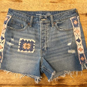 May include: Blue denim shorts with distressed details and crochet trim in white, pink, and blue. A crochet patch is sewn on the front. The shorts have a raw hem and the brand name "OLD NAVY" is visible.