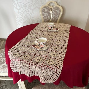 May include: A crocheted table runner in a cream color with a square pattern. The runner is laid on a red tablecloth and two white teacups with floral designs are placed on top of the runner.