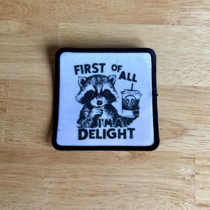 May include: A black and white patch with a raccoon holding a coffee cup. The text on the patch reads "First of all I'm a Delight."