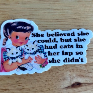May include: A sticker featuring a vintage illustration of a girl holding two kittens. The text on the sticker reads: "She believed she could, but she had cats in her lap so she didn't."