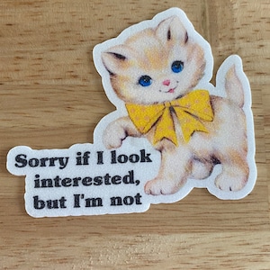 May include: A sticker featuring a vintage-style kitten with blue eyes and a yellow bow. The kitten has a slightly annoyed expression. Text on the sticker reads, "Sorry if I look interested, but I'm not."