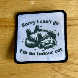 May include: White patch with black border featuring a black cat with green eyes and the text "Sorry I can't go I'm an indoor cat."