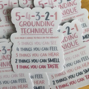 May include: A die-cut sticker featuring the 5-4-3-2-1 grounding technique in pink and dark gray text. The design lists: 5 things you can see, 4 things you can feel, 3 things you can hear, 2 things you can smell, and 1 thing you can taste.