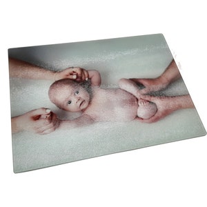Könnte beinhalten: Ein Schneidebrett aus Glas mit einem Foto eines Babys, das von zwei Händen gehalten wird. Das Baby liegt auf einer weißen Oberfläche und hat einen hellrosa Teint. Das Foto ist auf das Schneidebrett aus Glas gedruckt.