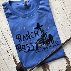 May include: A blue t-shirt with a black graphic design of a cowboy riding a horse. The graphic includes the text "The T-Roc Store Ranch Boss One Tough Cowgirl Brand".