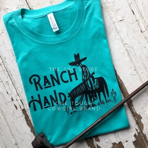 May include: Teal blue t-shirt with a black graphic of a cowboy riding a horse. The text "The T-Roc Store Ranch Hand One Tough Cowgirl Brand" is printed on the shirt.