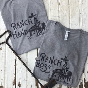 May include: Two gray t-shirts with black text and illustrations. The top shirt says "Ranch Hand" and features a cowboy riding a horse. The bottom shirt says "Ranch Boss" and features a cowboy riding a horse.
