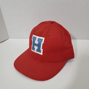 May include: A red baseball cap with a blue and white letter "H" patch on the front. The cap has a curved brim and a six-panel design. The hat is made of a durable fabric.