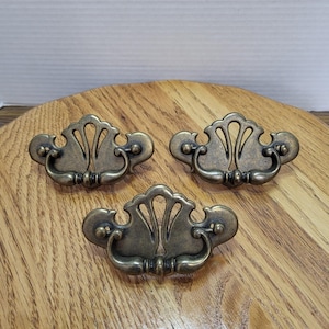 May include: Three matching drawer pulls made of antique brass-toned metal. Each pull features a decorative backplate with a fan-like cutout design and a curved, swinging handle attached to two circular mounting points on the base plate.