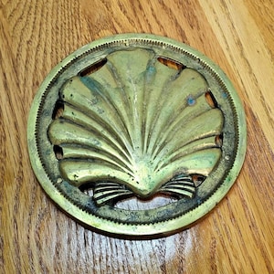 May include: A circular brass trivet featuring a central embossed scallop shell design. The metal surface displays a weathered patina with visible oxidation and a textured, serrated outer rim border. The piece has open cutouts surrounding the shell motif.
